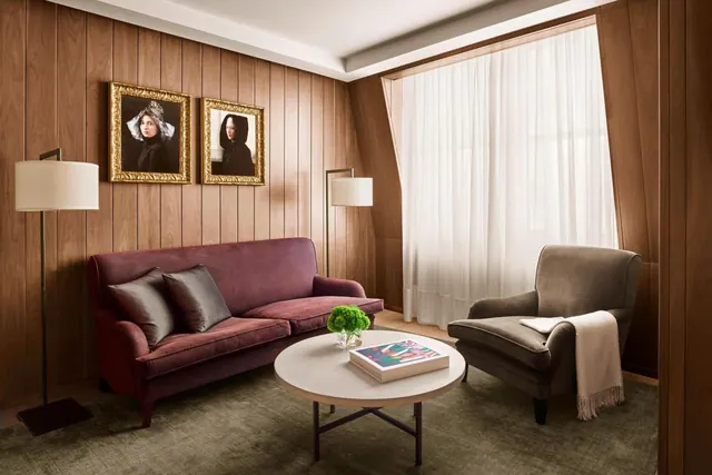 A living area with a burgundy velvet sofa, a grey armchair, a round coffee table and two framed portraits against wood-panelled walls.