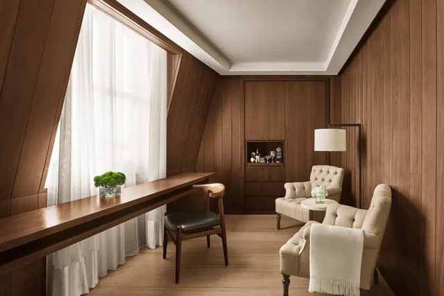 A wood-panelled sitting room with a long wooden desk by a sheer-curtained window, two tufted armchairs, a floor lamp and a built-in cabinet with glassware.
