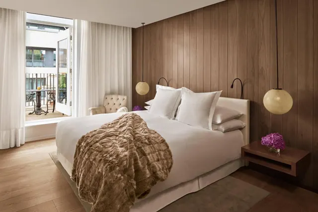 A bedroom with a made king bed under a fur throw, wood-panelled walls, pendant lights, and an open door onto a balcony with a table and chairs.