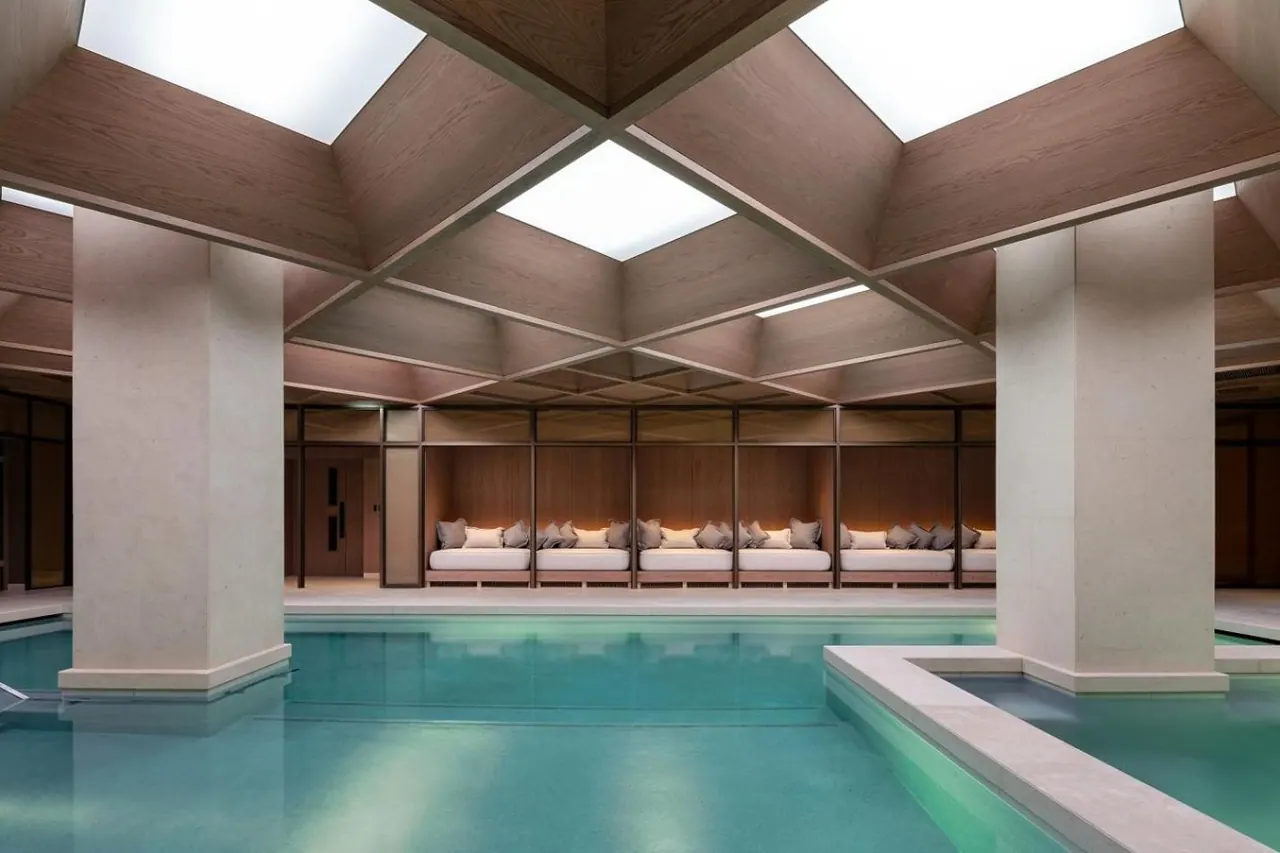 An indoor swimming pool under a geometric wooden ceiling with skylights, stone columns, and a row of cushioned daybeds along the back wall.