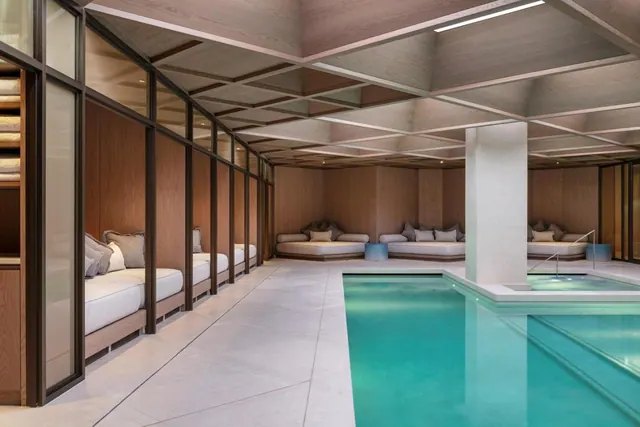 An indoor swimming pool with a small inset pool at right, lined by wood-panelled seating alcoves with cushioned daybeds.