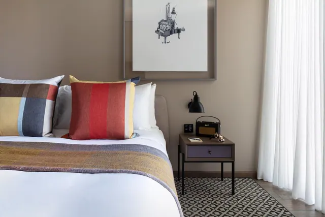 A made bed with striped and checked cushions sits beside a bedside table with a radio, a wall lamp and a framed print above.