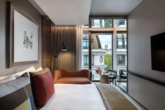 A bedroom with a made king bed, an orange armchair, a floor lamp, a small table with breakfast, and floor-to-ceiling windows overlooking neighbouring buildings.