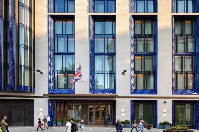 The front facade of The Londoner hotel with blue-panelled windows, a Union Jack flag, and pedestrians on the pavement below.
