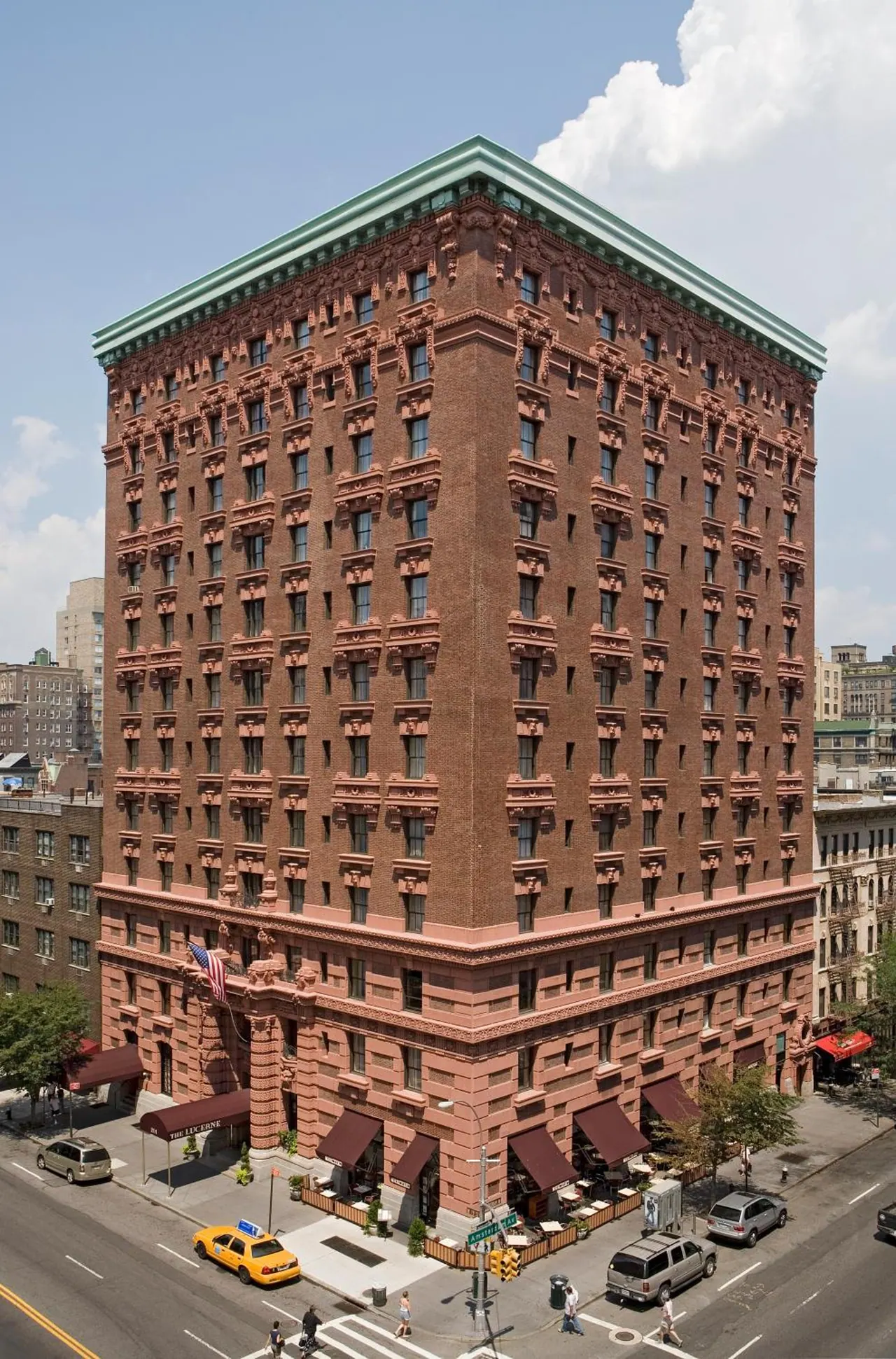 Exterior of a tall red brick hotel building with ornate stonework on a street corner, with sidewalk café tables under red awnings and a yellow taxi on the road below.