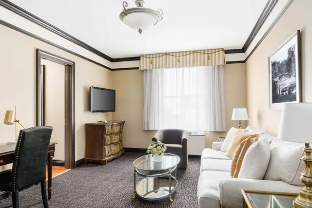 A hotel living area with a white sofa, glass coffee table, armchair, wall-mounted television, wooden dresser and a desk with a chair.