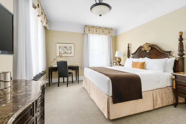 A bedroom with a king bed under a carved dark wood headboard, a writing desk and chair beneath a framed map, a marble-topped dresser, and a wall-mounted television on the left.