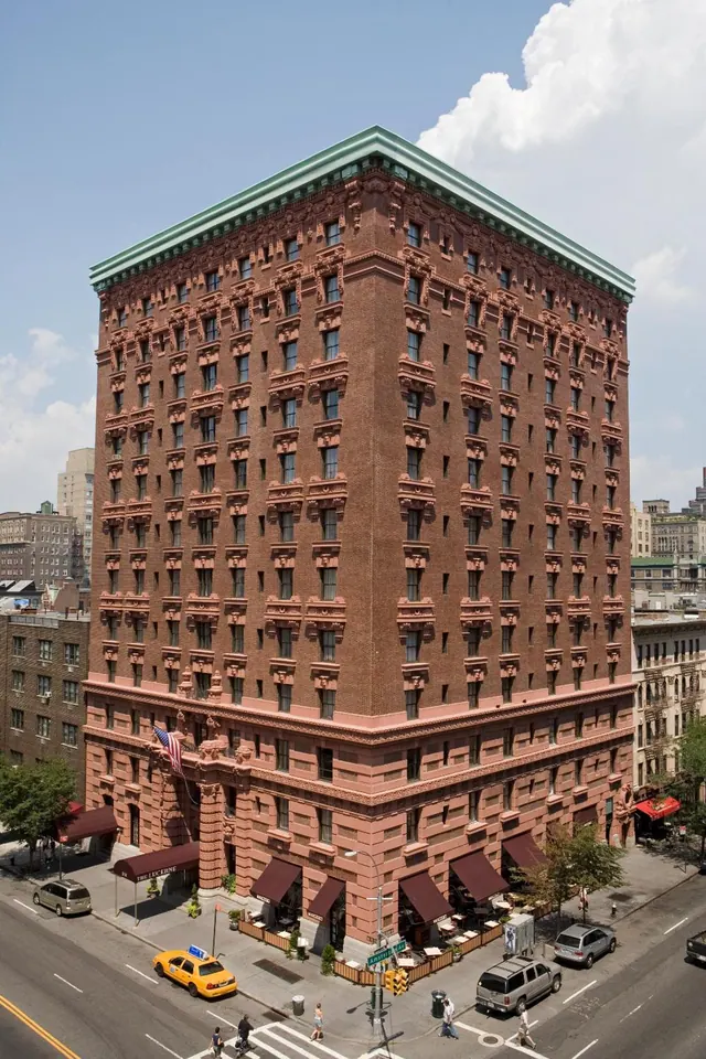 Exterior view of a red brick hotel building with terracotta ornamentation and a green copper roof, with a yellow taxi and outdoor dining tables at the street corner below.