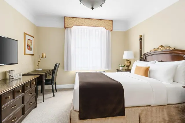 A bedroom with a king bed with a brown throw, a wooden desk and chair, a dresser with a wall-mounted television, and a window with sheer white curtains.