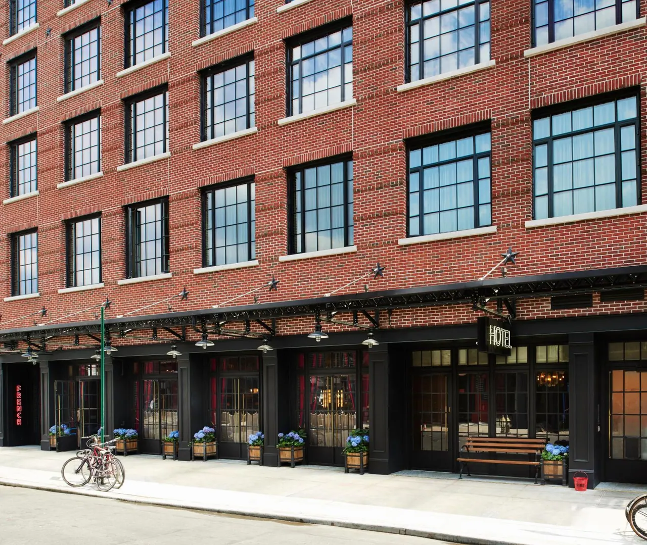 Brick facade of a hotel with a HOTEL sign above the entrance, planters with blue flowers, a wooden bench and parked bicycles on the sidewalk.