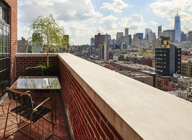 A brick-floored rooftop terrace with a glass table, two chairs and a small tree overlooks a city skyline that includes One World Trade Center.
