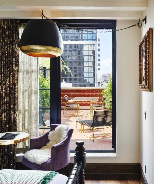 A bedroom with a purple armchair and sheepskin throw beside tall windows that open onto a brick-walled terrace with gold metal chairs and a table, with high-rise buildings beyond.