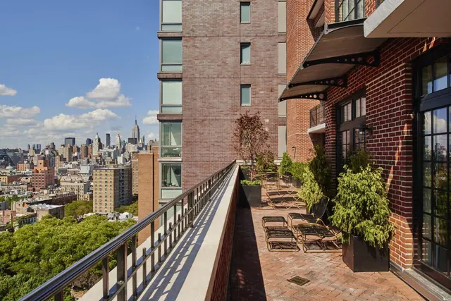 A brick-walled terrace with lounge chairs and potted shrubs overlooks a railing and a city skyline that includes the Empire State Building.