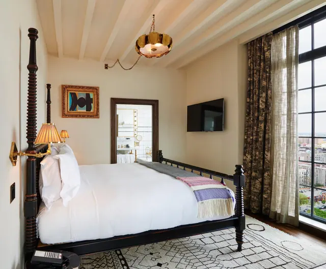 A bedroom with a dark wooden bed dressed in white linen, a wall-mounted television, and tall windows looking out over city buildings, with a bathroom visible through an open doorway.