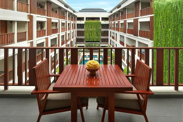A wooden table with two cushioned chairs and a yellow flower arrangement sits on a balcony overlooking a courtyard with a pool and a green plant wall.