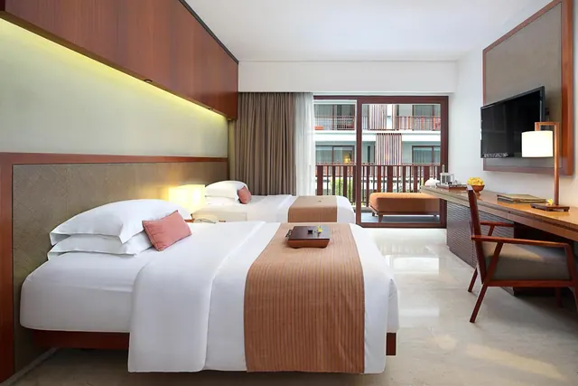 A twin-bedded hotel room with a wall-mounted television and desk, with a sliding door opening onto a balcony that has a cushioned daybed and a view of the neighbouring building.