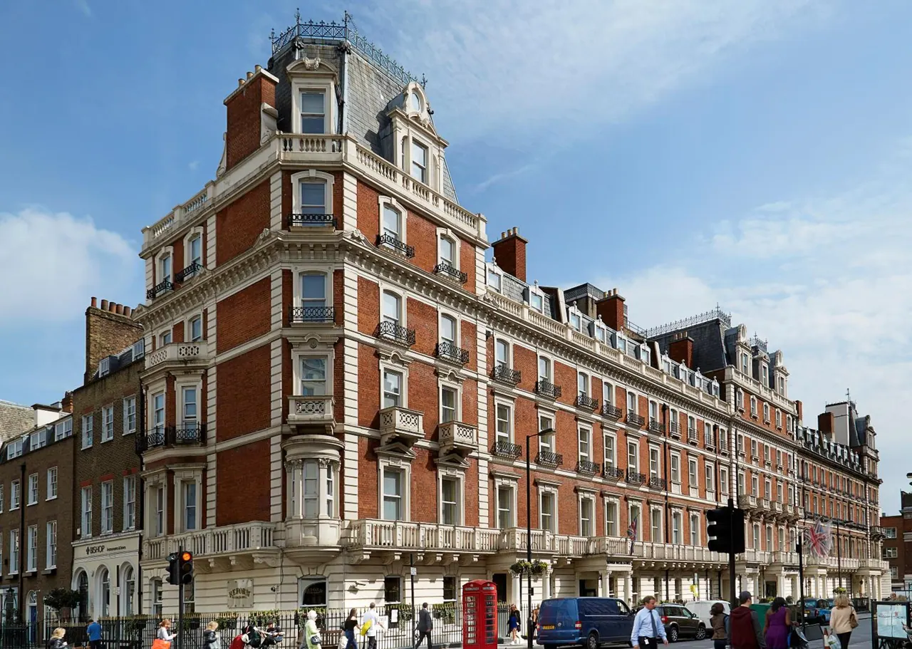 A large red-brick corner building with white stone trim and a mansard roof, with pedestrians, a red telephone box and a blue van on the street below.