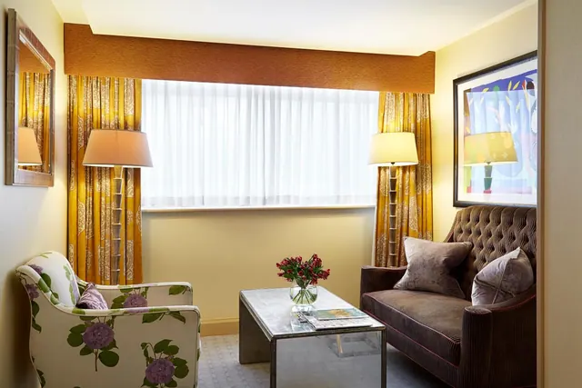 A seating area with a floral armchair, a brown tufted sofa, a glass coffee table holding a vase of flowers, and yellow curtains framing a window.