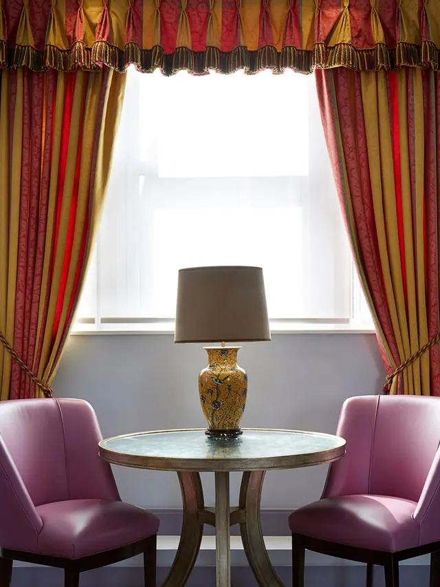 A small seating area with two pink leather armchairs, a round metal-topped table holding a yellow patterned lamp, and draped curtains at a window.