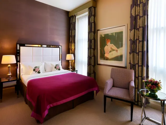 A hotel bedroom with a double bed under a magenta throw, two bedside lamps, an upholstered armchair and a framed painting on the wall.
