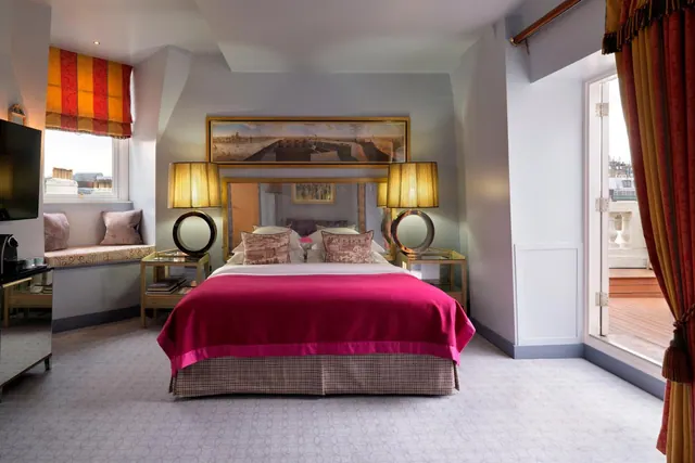 A bedroom with a made bed under a pink throw, bedside tables with lamps, a window seat, a wall-mounted television, and a glass door opening onto a terrace.