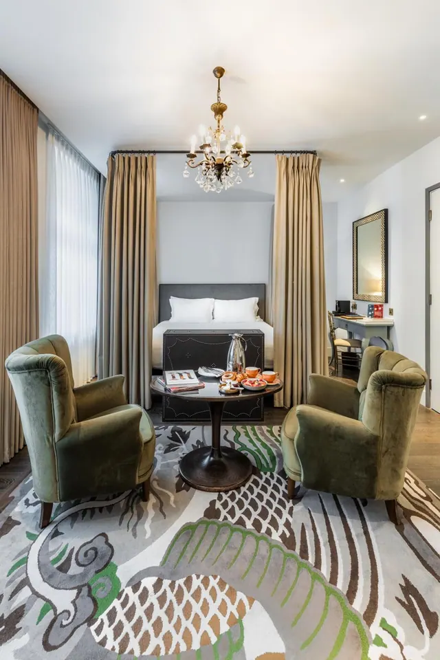 A hotel room with two green velvet armchairs around a round table holding breakfast items, with a bed in an alcove behind and a desk on the right.