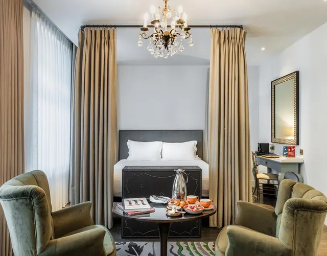 A hotel bedroom with a double bed between floor-length curtains, a crystal chandelier, two green velvet armchairs, a breakfast tray on a round table, and a desk with a mirror.