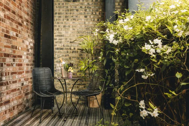 A small private balcony with two black wicker chairs, a round metal table holding a champagne bucket and glasses, brick walls and a flowering climbing plant.
