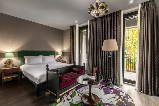 A bedroom with a king bed, a upholstered bench at the foot of the bed, a crystal chandelier, and a glass door opening onto a balcony with trees beyond.