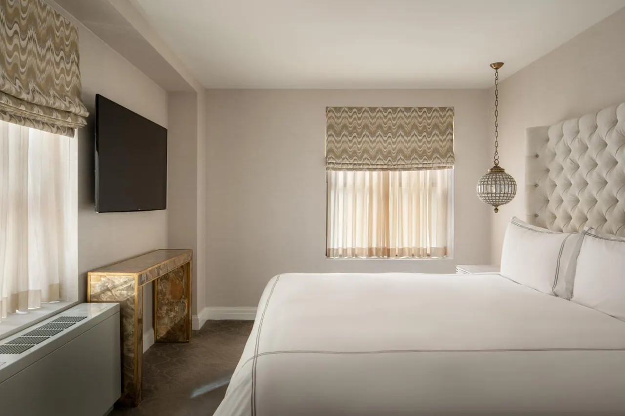 A bedroom with a made king bed, a wall-mounted flat-screen TV, a tufted headboard and a window with patterned roman shades.