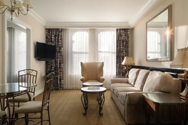 A living area with a beige sofa, an armchair, a round coffee table, a wall-mounted television and a dining table at the left.