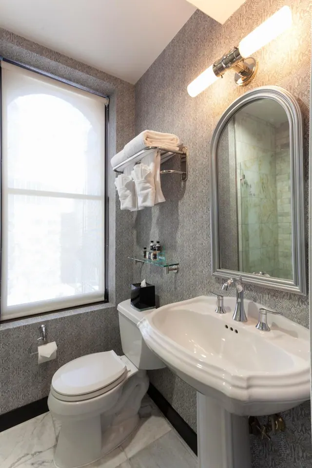 A bathroom with a pedestal sink, arched wall mirror, toilet, folded towels on a wall rack and a window with a roller blind.