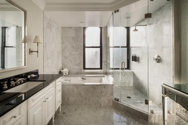 A marble-tiled bathroom with a drop-in bathtub beneath a frosted window, a glass-enclosed walk-in shower, and a black granite vanity with a single sink.