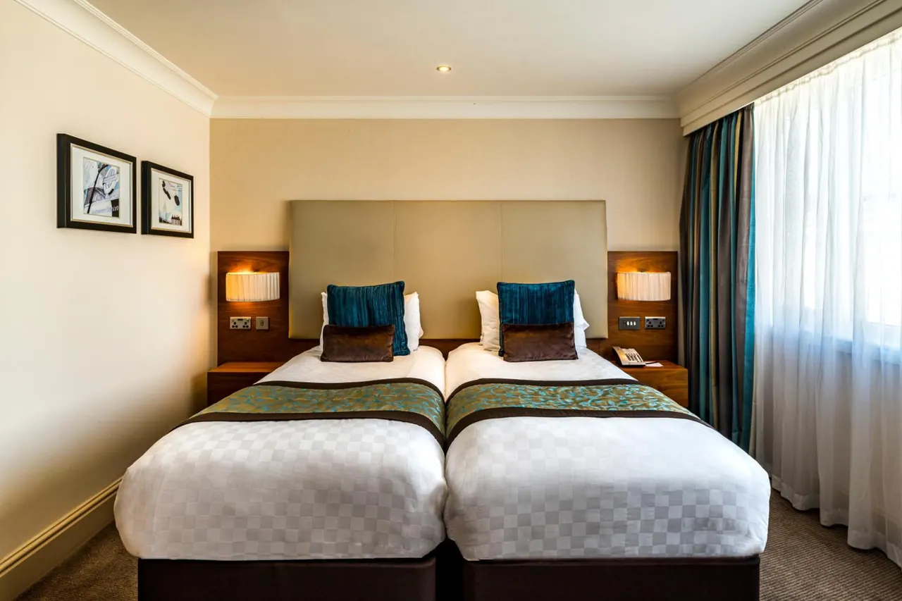 A twin bedroom with two made beds, teal and brown cushions, wall lamps on bedside tables, and sheer and striped curtains by the window.