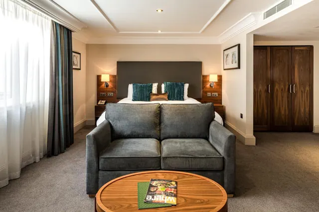 A hotel bedroom with a double bed behind a grey sofa and a wooden coffee table with magazines, with a wooden wardrobe on the right and curtained window on the left.
