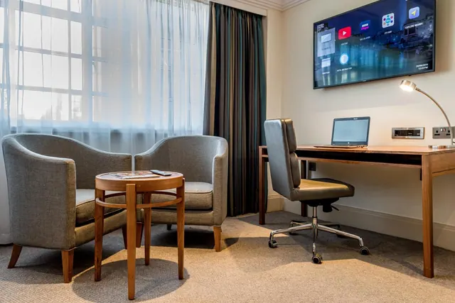 A seating area with two grey armchairs, a round wooden side table, a wooden desk with a laptop and office chair, and a wall-mounted television.