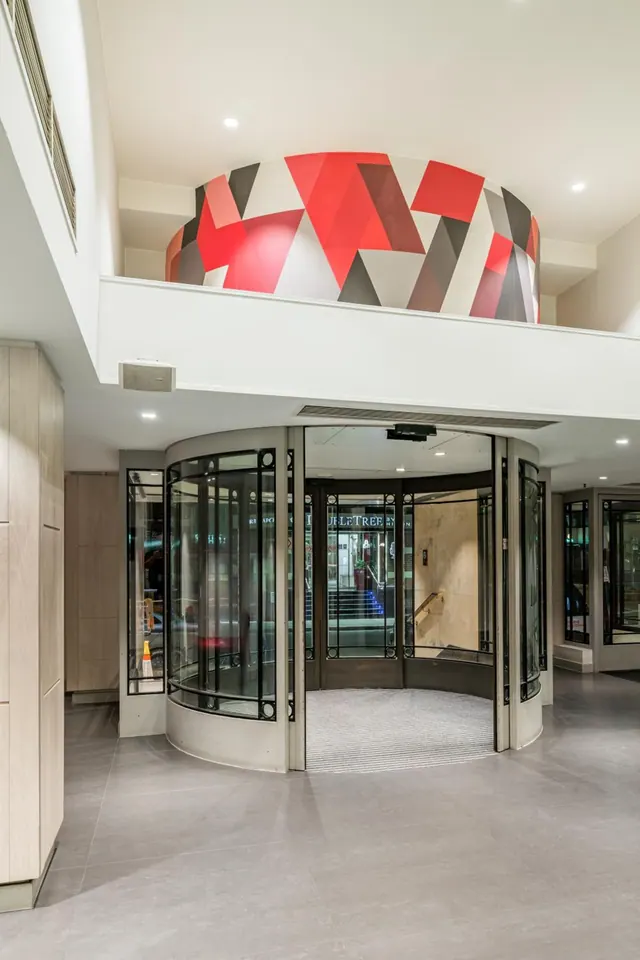 A revolving entrance door sits below a mezzanine with a red, grey and white geometric mural in a hotel lobby.