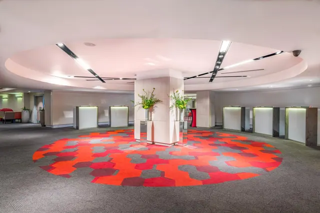 A large open foyer with a red and grey patterned carpet, a central pillar holding potted plants, and illuminated white counters along the walls under a circular ceiling.