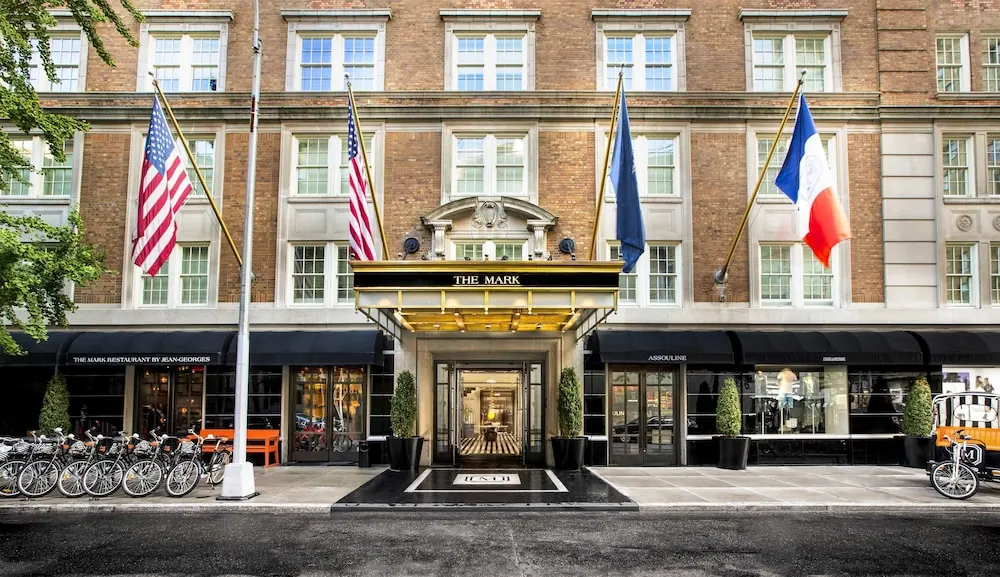 The front facade of The Mark hotel with its illuminated entrance canopy, three flags on poles, and a row of parked bicycles along the sidewalk.