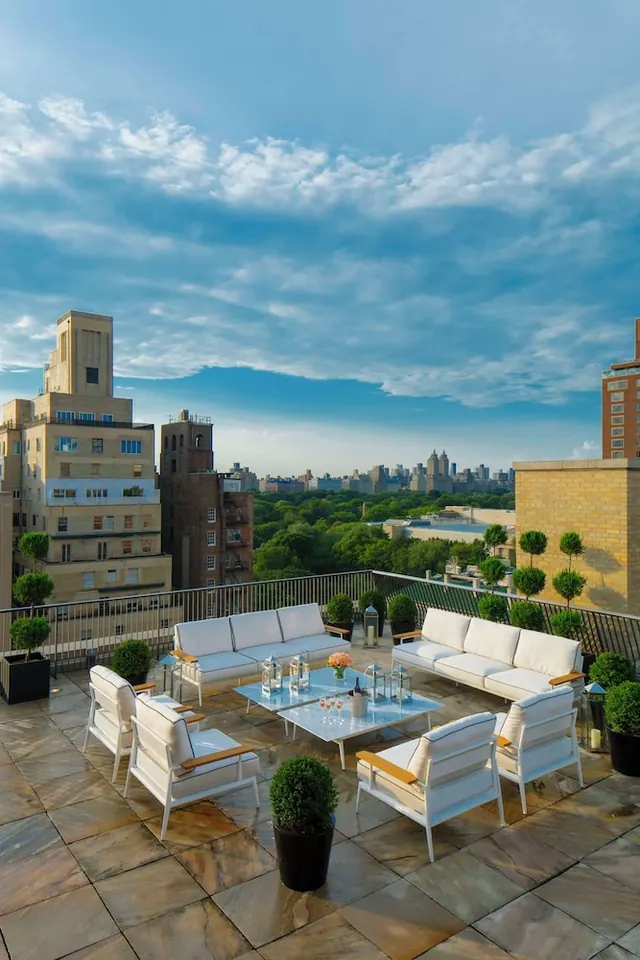 A rooftop terrace with white outdoor sofas, armchairs, glass-topped tables and potted topiary overlooks a city skyline and green park trees under a partly cloudy sky.