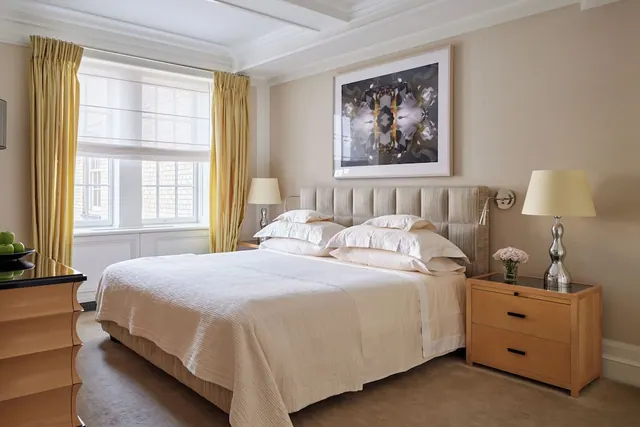 A bedroom with a king bed, upholstered headboard, two bedside lamps, a framed abstract artwork and yellow curtains at the window.