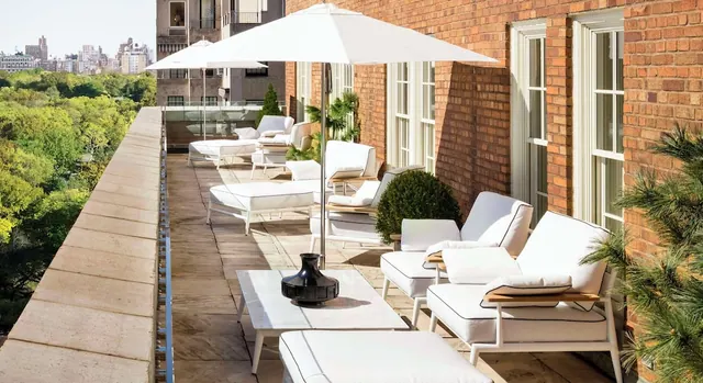 A brick-walled terrace with white lounge chairs, footstools, a side table and white umbrellas overlooks green trees and a city skyline.