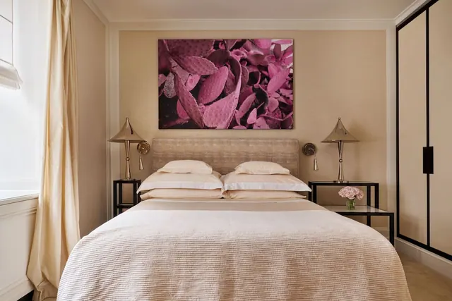 A bedroom with a made double bed, two metal bedside lamps, a pink botanical canvas on the wall and a tall wardrobe with black-framed doors on the right.