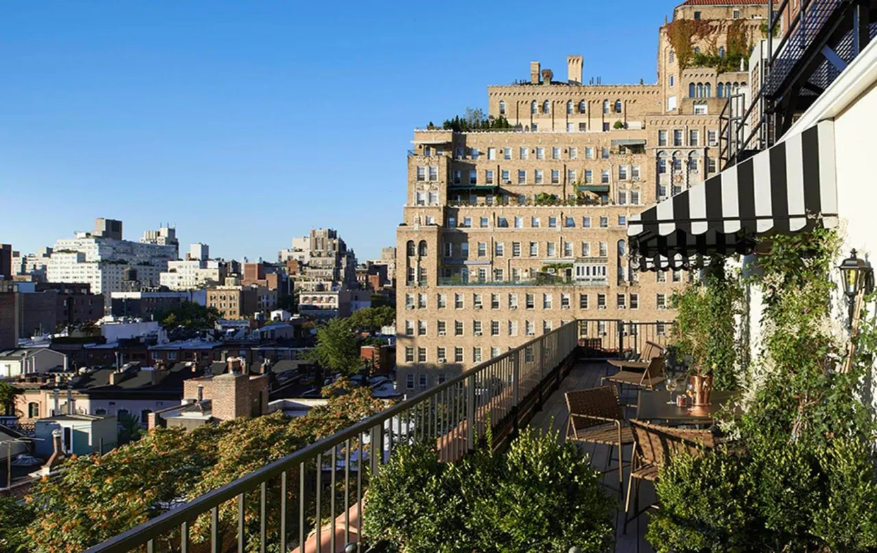 A rooftop terrace with wooden chairs, a small table, plants and a striped awning overlooks a brick apartment building and city rooftops.