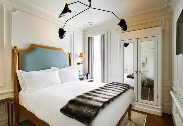 A bedroom with a made king bed under a black multi-arm ceiling light, a mirrored wardrobe, and a window with sheer curtains.