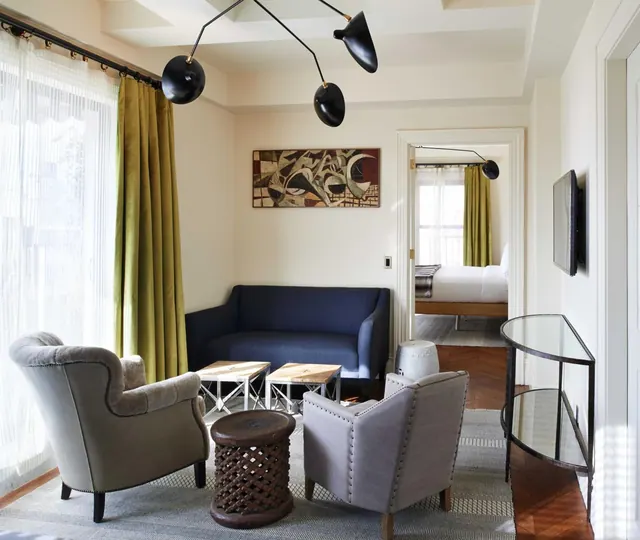 A living area with a navy sofa, two grey armchairs, wooden side tables and a bed visible through the open doorway to the bedroom.