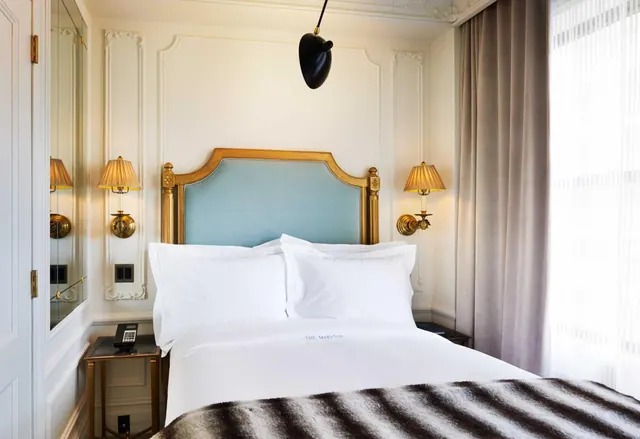 A made-up double bed with white linens and a gilded headboard with blue upholstery, flanked by wall lamps, beside a sheer-curtained window.