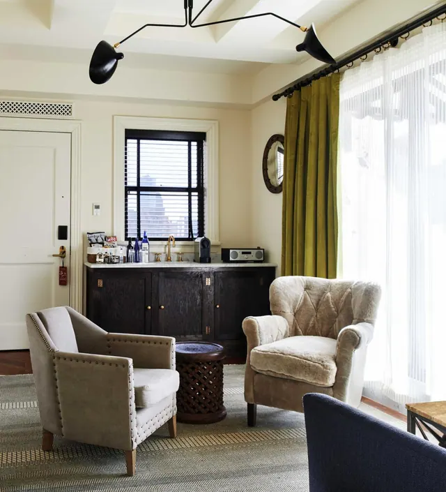 A hotel living area with two upholstered armchairs, a dark wooden sideboard with a sink and coffee machine, and sheer curtains beside green drapes at a tall window.