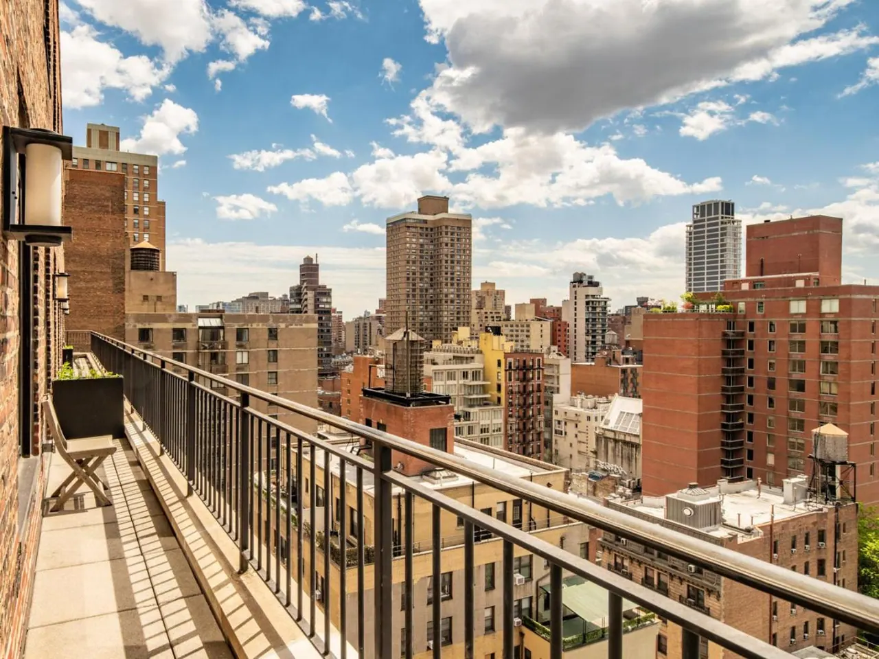 A private balcony with a metal railing, a wooden folding chair and a planter looks out over city rooftops and brick residential buildings under a partly cloudy sky.