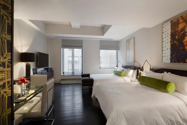 A bedroom with two made double beds with green bolster pillows, a glass-topped desk with chairs, a wall-mounted television, and two windows looking onto a building across the street.